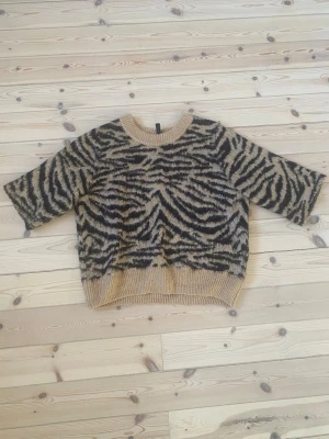 Beige zebramönstrad stickad tröja - Cool stickad tröja med zebramönster i beige och svart. Tröjan har rund hals, halvlånga ärmar och ribbade muddar vid hals, ärmslut och nederkant. Perfekt för dig som vill sticka ut med ett djurmönster i garderoben. Har klippt av ermarna men det syns inte 