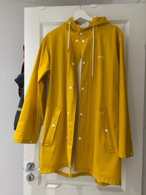 Gul regnjacka från Tretorn - Snygg gul regnjacka från Tretorn med huva och vita snören. Jackan har tryckknappar, två stora fickor framtill och är tillverkad i ett vattentätt material. Perfekt för regniga dagar och enkel att matcha med olika outfits.