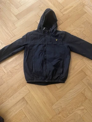 Svart vindjacka från Lyle & Scott - Snygg svart vindjacka från Lyle & Scott med huva och dragkedja framtill. Jackan har två fickor, elastisk nederkant och är prydd med den klassiska gula loggan på bröstet. Materialet är lätt och vindtätt, perfekt för blåsiga dagar.