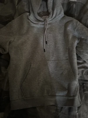 Grå hoodie från Jack & Jones - Klassisk grå hoodie från Jack & Jones med huva och dragsko. Tillverkad i mjukt sweatshirtmaterial och har en stor känguruficka framtill. Perfekt för en avslappnad och stilren stil.
