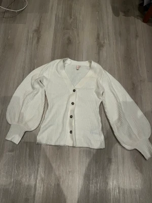 Vit kofta med puffärm från Nelly - Säljer en vit ribbstickad kofta från Nelly med stora puffärmar och v-ringning. Koftan har svarta knappar framtill och breda muddar vid ärmsluten. Använd endast 1 gång, storlek xxs💞