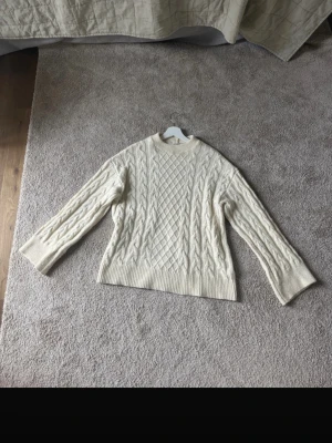 Krämvit kabelstickad tröja - Säljer en krämvit kabelstickad tröja med klassiskt mönster och rund hals. Tröjan har långa ärmar och breda ribbade muddar vid ärmslut och nederkant. Perfekt för lager-på-lager och riktigt mysig under kalla dagar.