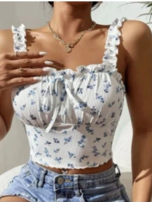 Blommig vit croppad linnetopp - Supersöt croppad linnetopp i vitt med blått blommönster och volangkant. Toppen har breda axelband med volangdetaljer och en liten rosett framtill. Perfekt för varma dagar och ger en romantisk vibe till din outfit.