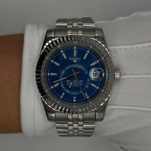 Seiko Sky Dweller blå urtavla - Stilren Seiko Sky Dweller klocka i rostfritt stål med blå urtavla och silverfärgade index. Klockan har datumfönster, räfflad bezel och länkarmband i metall. Automatisk rörelse syns genom glasbaksidan. Perfekt accessoar för dig som gillar klassisk men modern stil.