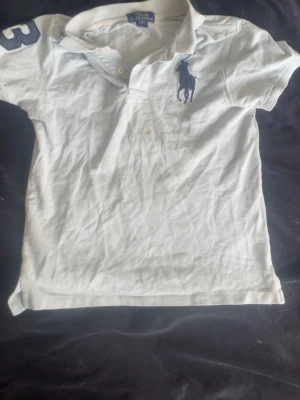 Ljusblå  pikétröja från Polo Ralph Lauren - Ljus blå pikétröja från Polo Ralph Lauren med klassisk krage👕, knappar och stor marinblå logga på bröstet. Tröjan har korta ärmar och en siffra på ena ärmen. Perfekt för en sportig och stilren look. Tveka inte att höra av dig vid några funderingar! Storlek M men sitter mer som S , Priset är ej hugget i sten 🏆