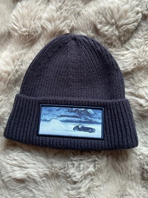 Mörkgrå ribbstickad mössa med patch - Snygg mörkgrå ribbstickad mössa med uppvikt kant och en patch framtill som visar ett vinterlandskap med en bil. Perfekt accessoar för kalla dagar och ger en cool touch till din outfit.