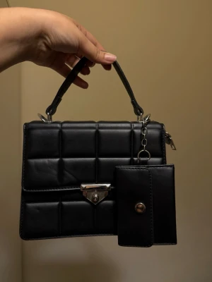 Svart quiltad handväska med plånbok - Stilren svart handväska i quiltad design med kort handtag och silverfärgat lås framtill. Väskan har en matchande svart plånbok som hänger i en kedja. Tillverkad i konstläder med rutmönster för en trendig look.