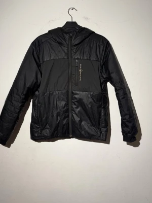 Svart vindjacka från Solid Racing - Snygg svart vindjacka från Solid Racing med huva och dragkedja framtill. Jackan har en bröstficka med dragkedja och logga, samt två sidofickor. Tillverkad i lätt, vattenavvisande material som passar perfekt för blåsiga dagar.