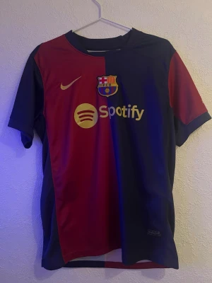 FC Barcelona matchtröja Nike - Säljer en officiell FC Barcelona matchtröja från Nike med klassisk röd och marinblå design. Tröjan har klubbmärke, Nike-logga och Spotify-sponsor framtill samt UNHCR-tryck bak. Korta ärmar och tillverkad i lätt polyester – perfekt för fotbollsträning eller match.