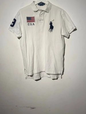Vit pikétröja från Ralph Lauren - Vit pikétröja från Ralph Lauren med klassisk krage och knappar. Stor USA-flagga och text på bröstet samt stor marinblå Polo-logga. Siffra på ärmen i mörkblått. Perfekt till en sportig och clean look.