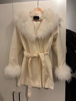 Vit pälsjacka med bälte från Garoff - Snygg vit pälsjacka från Garoff med stora fluffiga fuskpälskragar och muddar. Jackan har ett matchande midjebälte och en elegant, figurnära passform. Perfekt för dig som vill sticka ut i vinter och hålla dig varm med stil.