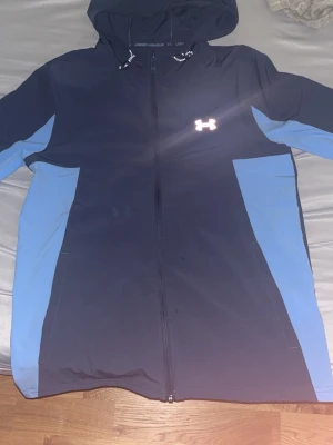 Blå och svart hoodie från Under Armour - Snygg hoodie från Under Armour i svart med blå paneler på sidorna och ärmarna. Dragkedja framtill, justerbar huva med snören och det klassiska Under Armour-loggan på bröstet. Perfekt för träning eller chill. Tillverkad i ett mjukt och stretchigt material.