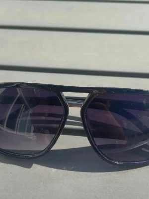 Solglasögon  - Snygga svarta aviator solglasögon med mörka glas och metallbåge. Klassisk pilotform med dubbla bryggor och silvriga detaljer vid sidorna. Perfekt accessoar för en cool och tidlös stil.