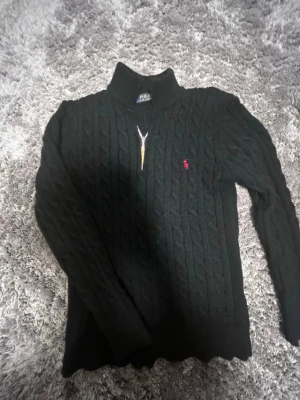 Svart kabelstickad tröja Polo Ralph Lauren - Kabelstickad svart tröja från Polo Ralph Lauren med röd broderad logga på bröstet. Tröjan har hög krage och långa ärmar. Klassisk och tidlös design i mjukt material som passar perfekt till jeans eller kjol.