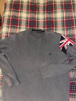 Grå stickad tröja från Henri Lloyd - Snygg grå stickad tröja från Henri Lloyd med rund halsringning och diskret logga broderad på bröstet. Tröjan har ribbade muddar och är tillverkad i mjukt material. Perfekt för lager-på-lager och enkel att matcha.