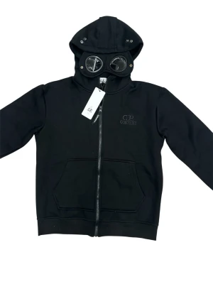 Svart hoodie med glasögon från C.P. Company- storlek M - Svart hoodie från C.P. Company med dragkedja, känguruficka och ikoniska glasögon i huvan. Tillverkad i mjukt bomullsmaterial och har diskret logga broderad på bröstet. Perfekt för dig som vill sticka ut med en unik streetwear-look. Storlek M