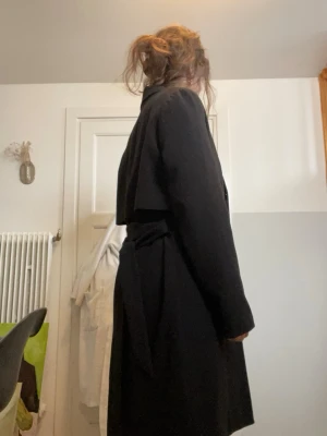 Svart kappa med knytskärp - Stilren svart kappa med klassisk krage och knytskärp i midjan. Jackan har en lös passform och ett extra lager tyg över ryggen för en cool look. Perfekt för dig som gillar minimalistisk stil. 