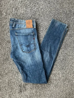Blå jeans från replay - Säljer ett par blå  jeans från replay med klassisk femficksdesign och snygga slitningar. Jeansen har en läderpatch bak i midjan och kontrastsömmar. Perfekta för dig som gillar en smal siluett och tidlös denimstil.