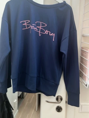 Blå sweatshirt från Björn Borg - Mörkblå sweatshirt från Björn Borg med rosa logotyp framtill. Tröjan har rund halsringning och långa ärmar. Perfekt för en avslappnad och sportig stil.