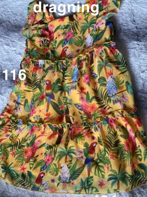 Gul klänning med papegojmönster - Färgglad gul klänning med tropiskt papegojmönster och gröna blad. Klänningen har volangdetaljer och är ärmlös med en axelbandslös design. Perfekt för varma dagar och somriga tillfällen.🌸 Dragningar i tyllen. Storlek 116