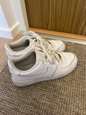 Vita Nike Air Force 1 sneakers - Klassiska vita Nike Air Force 1 sneakers i lågt snitt med ikonisk swoosh på sidan. Skorna har perforerad tå för ventilation, tjock sula och är tillverkade i läder med textilfoder. Perfekta för en clean och stilren look.