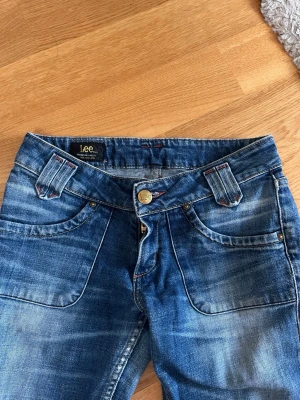 Blå jeans från Lee med detaljer - Snygga blå bootcut jeans från Lee med slitningar och coola detaljer som breda hällor och kontrastsömmar i rött. Klassisk femficksmodell med Lee-logga på knappar och bakficka. Passar mig bra, lite tajta och har vanligen strl 36. Bra längd på mig som är 163 cm