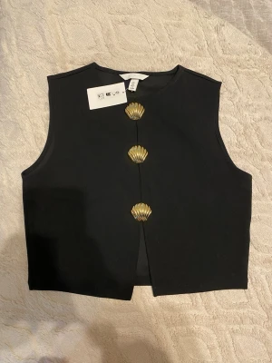 Svart väst med guldfärgade snäckknappar - Stilren svart väst från H&M med tre unika guldfärgade snäckformade knappar framtill. Västen är ärmlös, har rund halsringning och är tillverkad i ett mjukt tyg som ger en clean look. Perfekt för att lyfta vilken outfit som helst.