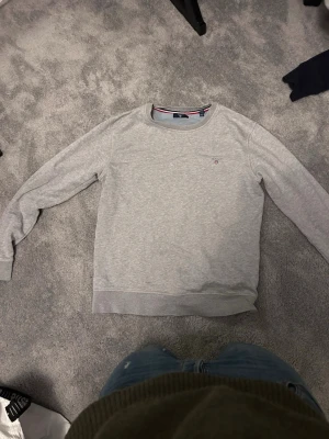 Grå sweatshirt från GANT - Klassisk grå sweatshirt från GANT med rund halsringning och diskret logga broderad på bröstet. Tröjan har ribbade muddar vid ärmslut och nederkant samt en snygg detalj med röd och marinblå rand på insidan av kragen. Perfekt för en clean och stilren look. Pris kan diskuteras mvh Felix⭐️ 