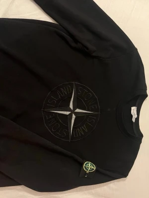 Svart Stone Island sweatshirt - Svart sweatshirt från Stone Island med stort broderat logotypmotiv på bröstet och klassisk patch på vänster ärm. Tröjan har rund halsringning och ribbade muddar. Perfekt för en stilren och trendig look. Pris kan diskuteras! Storlek L men funkar för M 