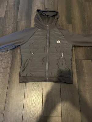 Mörkgrå hoodie från Moncler - Snygg mörkgrå hoodie från Moncler med quiltad front och två dragkedjeförsedda fickor. Klassisk huva och Moncler-logga på bröstet. Tillverkad i en mix av mjukt tyg och vadderad front för extra stil och komfort. Perfekt för dig som vill ha en exklusiv streetwear-look. Dragkedjan funkar men den saken har ramlat av där av priset så lågt 