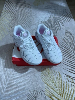 Nike Air Max Plus vita sneakers - Nike Air Max Plus sneakers i vitt mesh med detaljer i rosa och silver. Skorna har synlig Air Max-dämpning i sulan och klassisk swoosh-logga på sidan. Lågt skaft och snörning framtill. Perfekta för dig som gillar sportig och clean stil.