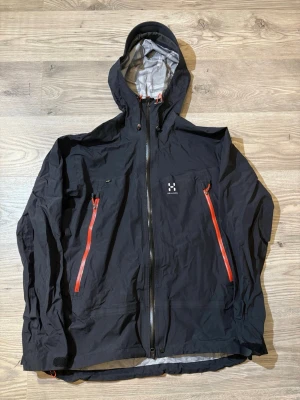 Haglöfs - Snygg svart regnjacka från Haglöfs med två röda dragkedjor på bröstfickorna och justerbar huva. Jackan har vattentätt material och hel dragkedja framtill. Perfekt för regniga dagar och outdoor-äventyr.