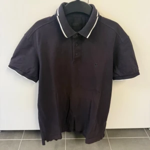 Svart pikétröja med vit kant Z Zegna - Klassisk svart pikétröja från Z Zegna med vit kant på krage och ärmslut. Tröjan har korta ärmar, knappar framtill och en liten logga på bröstet. Perfekt till jeans eller shorts för en stilren look.