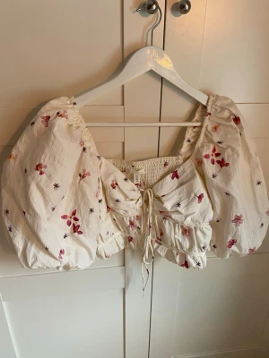 Blommig croppad blus med puffärmar - Supersöt croppad blus från H&M med blommigt mönster i rosa och rött. Toppen har stora puffärmar, snörning framtill och resår i midjan. Perfekt för dig som vill sticka ut med en romantisk och trendig look.
