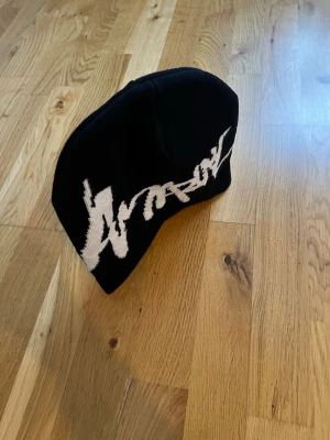 Svart  mössa med vit logga - Säljer en svart mössa med stor vit logga i graffiti-stil över framsidan. Mössan är ribbstickad och har en klassisk, rundad form. Perfekt för dig som vill ha en streetwear vibe och gillar tydliga prints. Helt ny