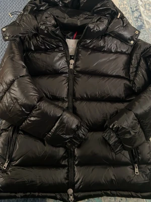 Svart dunjacka från Moncler - Snygg svart dunjacka från Moncler med glansig finish och vadderad design. Jackan har tvåvägsdragkedja, fickor med dragkedja och en hög krage med huva. Tillverkad i slitstarkt material och fylld med dun för extra värme. Perfekt för kalla vinterdagar.
