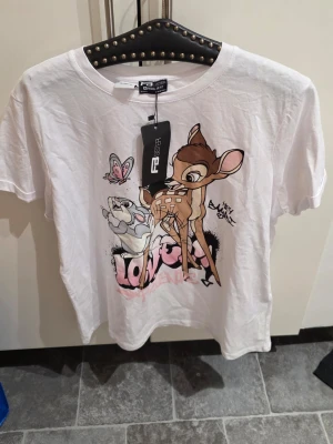 Vit Bambi t-shirt med tryck - Vit t-shirt från FB Sister med stort färgglatt tryck av Bambi och Stampe framtill. T-shirten har rund halsringning och korta ärmar. Materialet är mjukt och skönt, perfekt för en avslappnad stil.