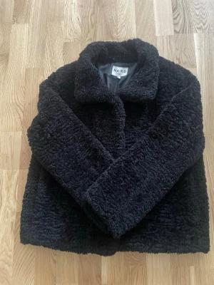 Svart teddyjacka från NA-KD - Mysig svart teddyjacka från NA-KD med klassisk krage och mjukt, fluffigt material. Jackan har rak passform och är helfodrad för extra komfort. Perfekt för att hålla värmen under kyliga dagar och ger en trendig look.