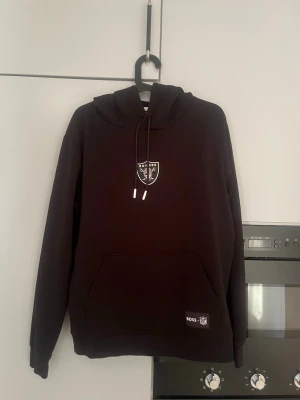 BOSS Raiders hoodie   - Snygg svart hoodie från BOSS med Raiders-logga på bröstet och stor RAIDER-text på ryggen. Klassisk känguruficka framtill, justerbar huva med snören och mjukt material. Perfekt för dig som gillar sportig streetstyle och vill sticka ut.