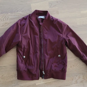 Bomberjacka med volangdetalj - Snygg vinröd bomberjacka från Ü i glansig polyester. Jackan har dragkedja framtill, ribbade muddar och coola volangdetaljer på ryggen och längs ärmarna. Perfekt för dig som vill sticka ut med en unik touch på klassisk streetstyle.