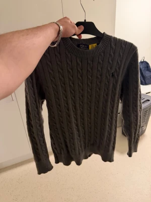 Mörkgrå kabelstickad tröja Polo Ralph Lauren - Säljer en HELT NY! mörkgrå kabelstickad tröja från Polo Ralph Lauren. Tröjan har rund halsringning, långa ärmar och klassiskt kabelmönster. Perfekt för dig som gillar stilrena och tidlösa plagg med en snygg passform Priset är ej hugget i sten!.