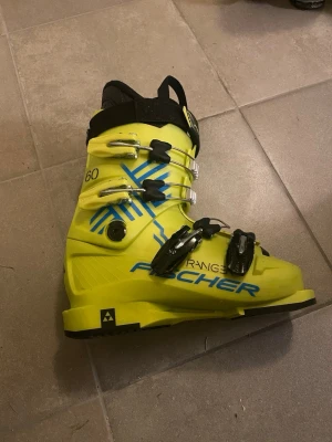 Gula Fischer pjäxor med spännen - Säljer ett par riktigt coola gula Fischer Ranger 60 pjäxor med blå detaljer och fyra metallspännen. Insidan är svart och de har en rejäl kardborre upptill för extra stöd. Perfekta för dig som vill sticka ut i backen! Storlek 23,5 och 273 mm