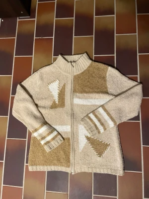 Beige stickad kofta från New Sensation - Sjukt unik stickad kofta från New Sensation med dragkedja framtill. Koftan har grafiska detaljer i vitt och brunt, ribbad krage och muddar samt vita och bruna ränder på ärmarna. Perfekt för lager på lager under kyliga dagar. Dragkedjan sitter lite löst som syns på sista bilden 