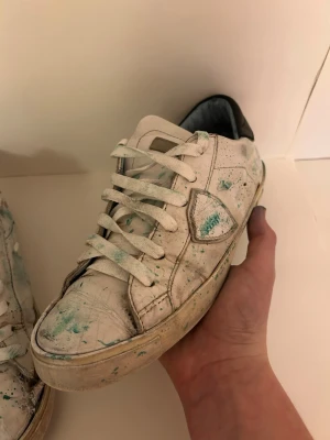  Custom Philippe model skor - Säljer ett par vita Philippe Model sneakers i skinn med gröna färgstänk och svart detalj på hälen. Skorna har klassisk snörning och låg profil. Perfekta för dig som gillar en unik och kreativ look.