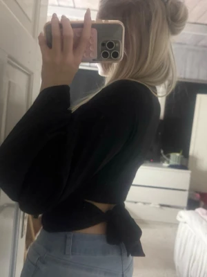 Svart croppad topp med knyt i ryggen - Säljer en svart långärmad croppad topp med snygg knytning i ryggen. Toppen har en tight passform och är perfekt att styla med jeans eller kjol. Enkel och stilren design som passar till många olika looks.