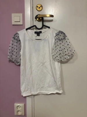 Vit topp med puffärmar från Primark - Säljer en vit topp från Primark i storlek S. Toppen har transparenta puffärmar i mesh med svarta prickar som ger en unik look. Kroppen är i mjuk bomull och modellen är normalpassad. Perfekt för dig som vill ha något extra till din outfit.