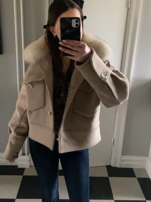 Beige ulljacka med stor krage - Säljer en beige jacka med stor fluffig krage och rejäla fickor framtill. Jackan har knappar och är perfekt för dig som vill ha en snygg och trendig look under vår och vinter. St S/M