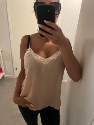 linne med spetskant - Säljer ett linne i blankare material för en mer exklusiv känsla med tunna axelband och v-ringning dekorerad med vit spets. Toppen har en lös passform och är tillverkad i ett slätt, mjukt material som ger en lyxig känsla. Perfekt för dig som gillar en enkel men ändå söt stil. Ca men passar även s