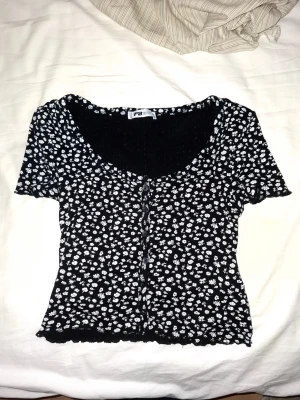 Coquette svart topp  - Kortärmad topp från FB Sister i svart med små vita blommor över hela tyget. Modellen har knappar framtill och lätt croppad passform. Tillverkad i mjuk bomullsmix som känns skön mot huden. Perfekt för dig som gillar en söt och enkel stil. Polkadot liknande blommor. 