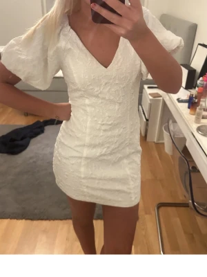 Vit kort klänning med puffärm - Supersöt vit kort klänning med puffiga kortärmar och v-ringning. Klänningen har en strukturerad yta som ger en lyxig känsla och sitter figurnära. Perfekt för dig som vill ha en stilren och trendig look.
